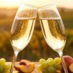 Duas taças de espumante brindando com uvas moscatel e pêssegos diante de um vinhedo ao entardecer