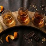 Cinco copos de whisky em tons de âmbar sobre aduela de barril, com casca de laranja e chocolate amargo, vistos de cima